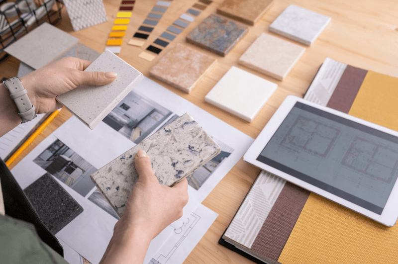 Close-up of a young female designer’s hands holding two marble tile samples over a wooden table, with a digital tablet and printed photos of home interiors used for design planning.